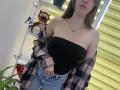 Nesie with bare shoulders, black strapless top, plaid shirt off, blue jeans, necklace, and long brown hair, standing near a tiger stuffed animal and butterfly decorations.