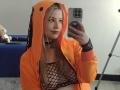 Melani Becker met blond haar, een oranje hooded jas, een nethemdje, waarbij haar borsten zichtbaar zijn, die een selfie maakt, en een zwarte choker draagt, in een kamer met een blauwe bank en een pluchen speelgoed..