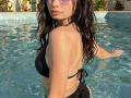 VikkiRi with wet black bikini, small tits, and long dark hair in a pool, wearing sunglasses, looking over shoulder, water droplets on skin, hands in water.