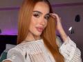 MarissaFex has long, straight orange hair, full pink lips, and a white lace top. She's touching her hair with a tattoo on her left hand.
