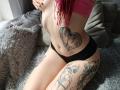 RachelNervis with red hair, pink top, black panties, tattoos on belly, thigh, and leg, sitting on gray fuzzy bed, hand on her ass, sunlight from window.