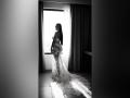 MiilaWx stands in a black-and-white photo, wearing a lace dress with a long, flowing train, showing her ass and back, looking over her shoulder at a window.