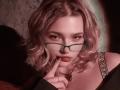 AdrianePittillo with wavy blonde hair, green glasses, and a finger touching her lips, wearing a black off-shoulder top with a jeweled strap, looking sultry.