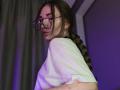 LelaTraverso with glasses, long brown braid, white tee, puffy red skin on arm, side profile, subtle smile, purple lighting.
