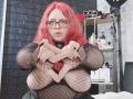 OnixDamon with red hair, glasses, and fishnet top, showing off large tits with her hands, in a black-tiled room, holding a dildo.