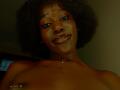 Close-up of KharlaQueen with dark brown skin, curly black hair, and a small, confident smile. She has full lips with dark red lipstick and is topless, showing a small tattoo on her right shoulder. A thin gold necklace with a small pendant is around her neck. The light is dim, casting a warm, shadowy glow on her face and upper body. The angle is slightly low, emphasizing her facial features and upper torso. Her eyes are dark and slightly upturned, adding to her confident expression.