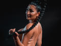 ItsJessieBabe mit glatter Haut, geflochtenem schwarzem Haar, glitzernden Gesichtsschmuck, die ihre lange Frisur hält, oben ohne, die Brüste sichtbar, dunkler Hintergrund, dezente Tattoos auf dem Rücken..