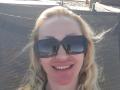 MysteryLaddy has blonde hair, blue sunglasses, and a red sunburn on her nose, smiling with white teeth. Sunlight reflects off her glasses.