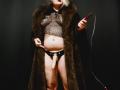 BeccaVanBoa with a fake cock in black mesh top, brown fur coat, and black panties, holding a red flogger. Her tits are visible through the mesh.
