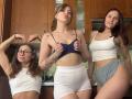 Three HotCaramelsTrio standing in a kitchen, wearing crop tops and high-waisted shorts. Center HotCaramelsTrio has small tits, lifting a blue top, showing tattoos. Left and right HotCaramelsTrio smile.
