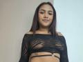 AmyMias with medium brown skin, black lace top showing her tits, straight brown hair, subtle makeup, small tattoo on right side, and a calm smile, standing in front.