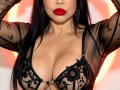Angelina Deleon mit roten Lippen, schwarzer Spitzen-BH, der große Brüste zeigt, ein durchsichtiges schwarzes Oberteil mit floralen Mustern, Arme erhoben, langes schwarzes Haar, goldene Ohrringe, gebräunte Haut, sexy..