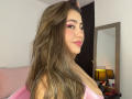 LorensDouce with long brown hair, pink eyeshadow, and pink lipstick, wearing a pink top, showing her tits, smiling, and looking over her shoulder in a bedroom.