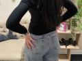 Dalaila's black long-sleeve top, blue jeans, and her hand on her ass, showing her butt. Long dark hair, silver bracelet, beige couch, and wooden shelf with green plant.