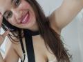SanetPerlas with long brown hair, smiling with pink lipstick, wearing a black strap halter top, showing her tits, in a selfie, raising her right arm.