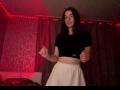 SunnyKlusman with glasses, black crop top, and white skirt, standing in dim room with red LED lights, mid-motion, hands up, brown hair, slight smile.