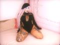 Chili, with pale skin and pink-bleached hair, kneels on a bed wearing a black fishnet bodysuit, showing large tattoos on her thighs, and wearing a black beret.