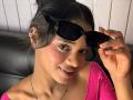 KiaraBrown with dark hair in a bun, wearing black sunglasses, pink sparkly top, and gold earrings, smiling with flushed cheeks, holding sunglasses with her right hand.