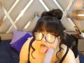 LillyMayer with black hair in a bun, glasses, tongue out, sucking a large, pink, erect dildo. She's wearing a black, sparkly top, and is on a bed.