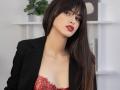 JasmineDay with long brown hair, red lipstick, black blazer, and red lace bra, showing cleavage, looking slightly to the side, with a serious expression.