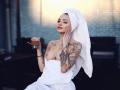KayaBelle with a white towel wrapped around her lower body, holding a cup of coffee, showing her tits, tattoos on her arms and chest, and a towel on her head.