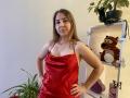Aminas with medium tits in a shiny red satin dress, hands on hips, smiling slightly, standing near a shelf with a tiger stuffed toy and green plant.