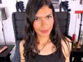 LunarisSmith, com cabelos pretos longos, olhos castanhos e um leve sorriso, vestindo uma blusa preta, tem um lábio com piercing e um colar, sentada em uma cadeira de couro preto, com um brinquedo sexual..