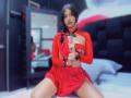 MelisaDaltonn in red outfit with cutout chest, holding a strap, sitting with legs apart, wearing black cuffs, looking at camera, bedroom, mirror reflecting her, medium skin, dark hair.