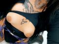 DenyChantal's big tits are exposed, with dark nipples, in a black top, showing tattoos on her chest and arms. She's wearing purple belts and has black hair.