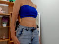 Steisii with a blue strapless top, showing her flat stomach and small ass, wearing high-waisted, light blue jeans, tattoo on left arm, and a necklace, in a room.