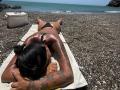 MajoJones, lying face down on a towel, sunbathing on a rocky beach, wearing a black thong bikini, with glistening oil-covered skin, and visible tattoos on her arm.
