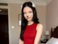 CherelleGess with black hair, red sleeveless top, white bow hair clips, fake black cat whiskers and nose, standing in a bedroom with a wooden door and lamp.