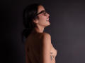 JennTylor with dark hair, glasses, smiling, topless, showing small tits, tattoo on arm, back to camera, dark background, side profile, looking right, wearing earrings.