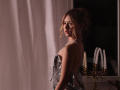 RosaVance with wavy brown hair, wearing a sparkly silver dress and pearl necklace, looking back over her shoulder, standing in a dimly lit room with white candles.