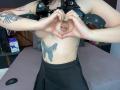 GoddessLuxliv making a heart shape with her hands over her small, pierced tit. She's got tattoos on her arm and belly, wearing a black leather top and skirt.