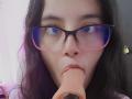 SamanthaBlow with glasses, nose ring, and dark hair, sucking a large, flesh-colored dildo. Her blue eyes are focused forward. Her lips are wrapped around the dildo.