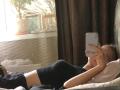 Hilaria Tabar est allongée sur un lit, vêtue d'un haut noir, et prend un selfie avec un téléphone blanc, tout en cachant son visage et en montrant ses fesses nues. On peut voir des rideaux marron en arrière-plan..