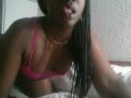 BettyBoopchi, dark-skinned, with long braids, pink bra, showing tits, gold necklace, tongue out, on bed, white sheets, textured wall in background.