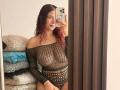 ValeryScot with red curly hair, wearing a black fishnet top, exposing her tits, standing in front of a mirror, holding a phone, with a confident smile.