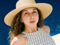 Sanella with wavy brown hair, wearing a white polka-dot dress and a straw hat, has a fair skin, small earrings, and a relaxed expression, against a deep blue background.