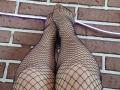Tinelia's legs in black fishnet stockings, feet pointed upwards, touching. Visible tan skin, purple LED strip along brick wall. Boobs not visible, focus on legs and feet.