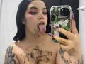 AryButty with chocolate smeared on her face, tongue out, holding a phone with green and black patterned case, long nails, tattoos on chest and arms, in a bathroom.