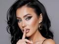 MistressTaylor with black wavy hair, blue eyes, and gold earrings, finger to lips, seductive look, shiny makeup, touching her finger to her slightly parted, glossy lips.