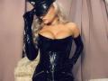 MademoiselleBella with blonde hair, wearing a black leather corset, gloves, and hat, has big tits, a choker, and is holding her hat, posing seductively.