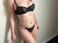 AlicePrings with pale skin, black sheer bra, and black lace thong, standing against a wall, showing her tits and pussy, with a small belly button piercing.