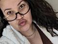 Brendabbew tiene labios gruesos, ojos marrones, cabello negro rizado y usa gafas con un piercing en la nariz. Lleva una chaqueta blanca y esponjosa y está mirando a la cámara..