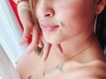 MirandaDreams with puffy lips, gold hoop earrings, and a butterfly tattoo on her chest, showing off her bare boobs with a hand near her face. She's wearing a gold necklace.