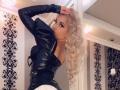 MademoiselleBella with long, curly blonde hair, black leather top, and white skirt, looking over her shoulder, hand in hair, in a stylish room.