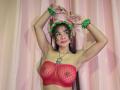 Yurimy with red sheer bralette, green flower headband, green wristbands, arms raised, pink curtain background, long dark hair, red lipstick, small tattoo above boobs, necklace with pendant.