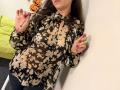 Tilliana, com cabelo castanho, vestindo uma blusa preta com estampas florais, segurando um barbante, com unhas cor-de-rosa, em pé em frente a uma parede branca, mostrando os seios, com uma cama verde ao fundo..